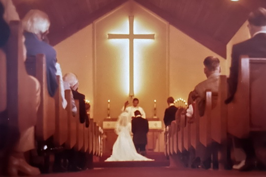 A man and woman receive the marriage blessing at the altar under a glowing cross.